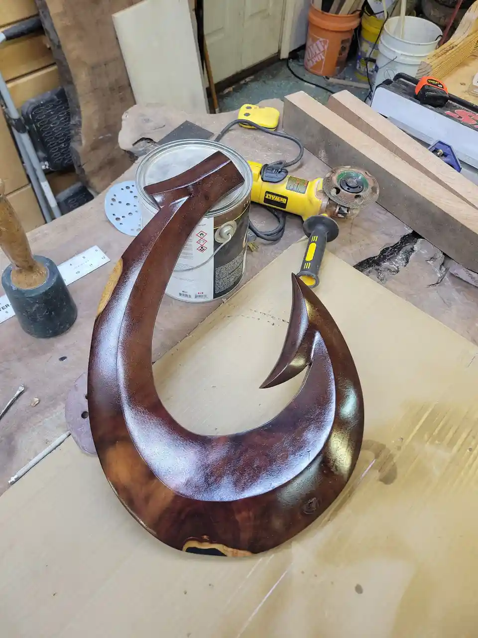 A large, polished wooden hook sculpture sits on a workbench surrounded by woodworking tools like a DeWalt angle grinder…