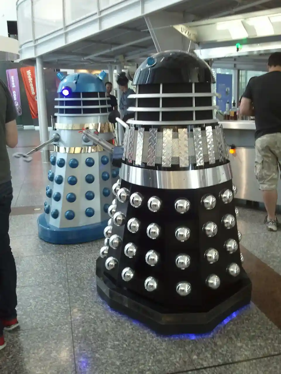 Two Dalek statues, one classic black and one blue, stand side by side in a convention hall. The black Dalek has overlaid…