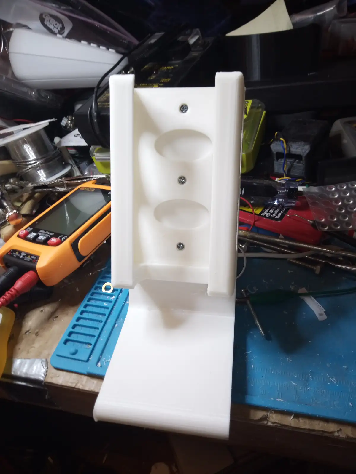 A white 3d-printed plastic object, shaped like a wall-mounted holder with two oval cutouts and three screws, stands upright…