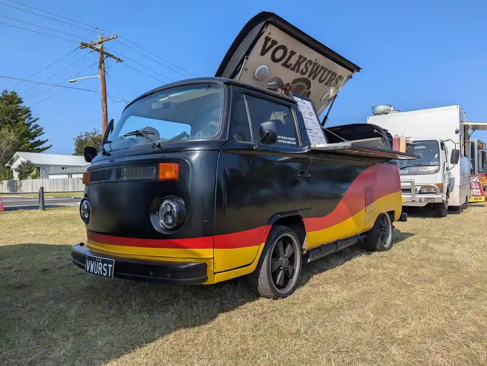 A black Volkswagen van with red, yellow, and black stripes resembling the German flag is parked on grass. The van has a…