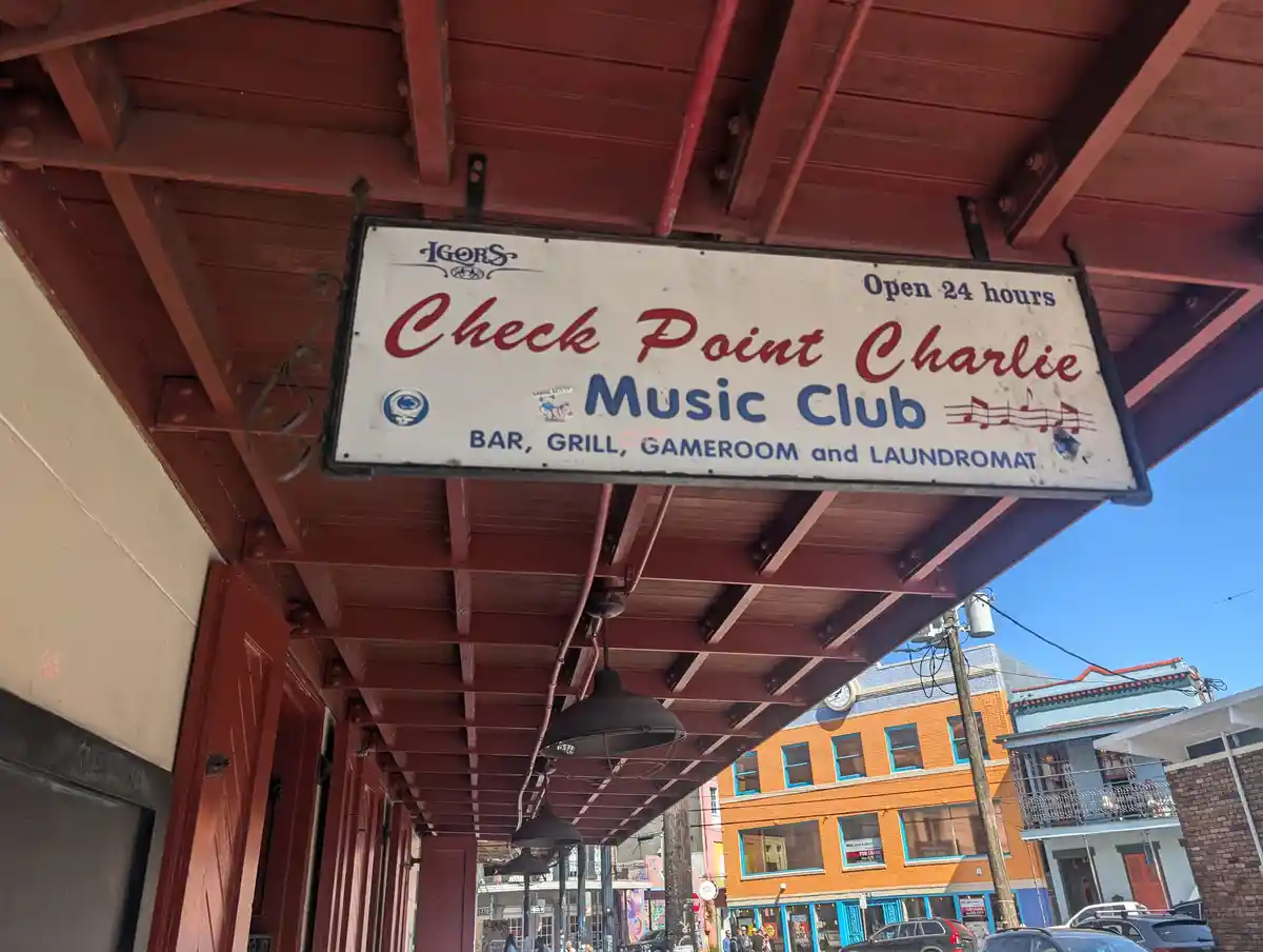 A white sign for 'Check Point Charlie Music Club' hangs under a red wooden awning, listing services as 'BAR, GRILL, GAMEROOM…
