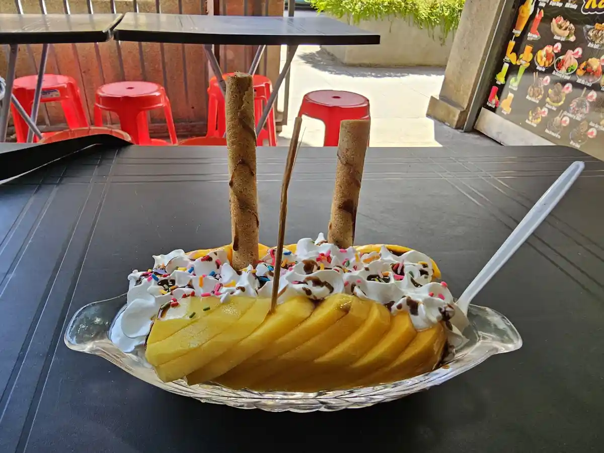 a dessert dish filled with sliced mango, whipped cream, chocolate syrup, and colorful sprinkles, with two wafer sticks…