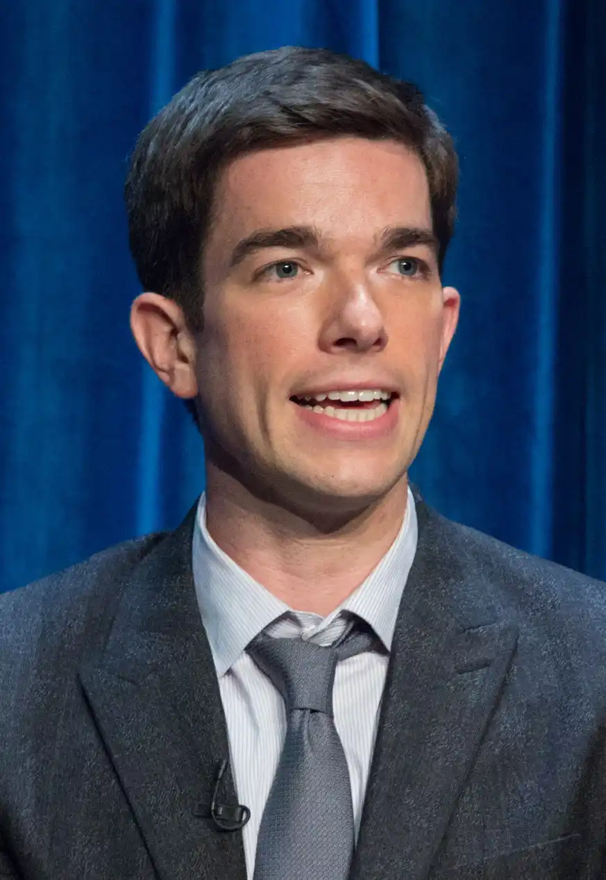a man in a gray suit and tie smiles while looking slightly upward, standing before a blue curtain. The overlaid text reads…