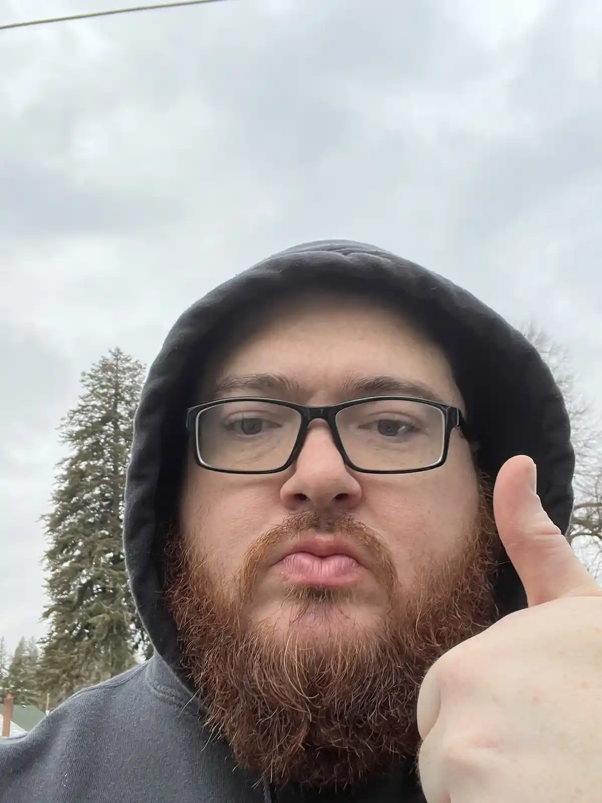 A man with a reddish beard and black-rimmed glasses, wearing a black hooded sweatshirt, gives a thumbs-up while making a…