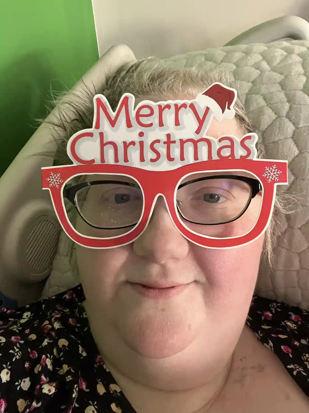 A person with short gray hair and glasses is sitting in a chair, wearing a red cardboard cutout of oversized glasses with…