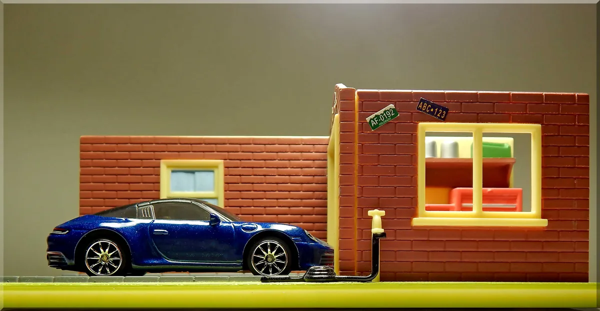 A blue toy sports car is parked beside a small toy brick building with a window showing furniture inside. Two toy license…