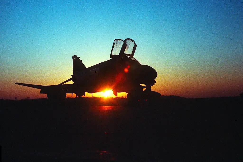 A silhouette of a fighter jet parked on a runway at sunset, with the sun directly behind it creating a bright lens flare…