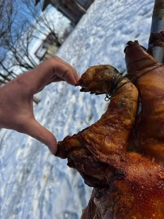 A person's hand forms a heart shape with their thumb and index finger near the head of a roasted pig hanging outdoors in…