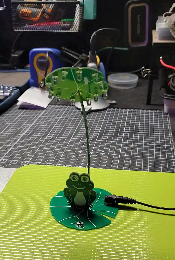 A handmade electronic project features a smiling green frog cutout sitting on a green lily pad base, with a thin green stem…