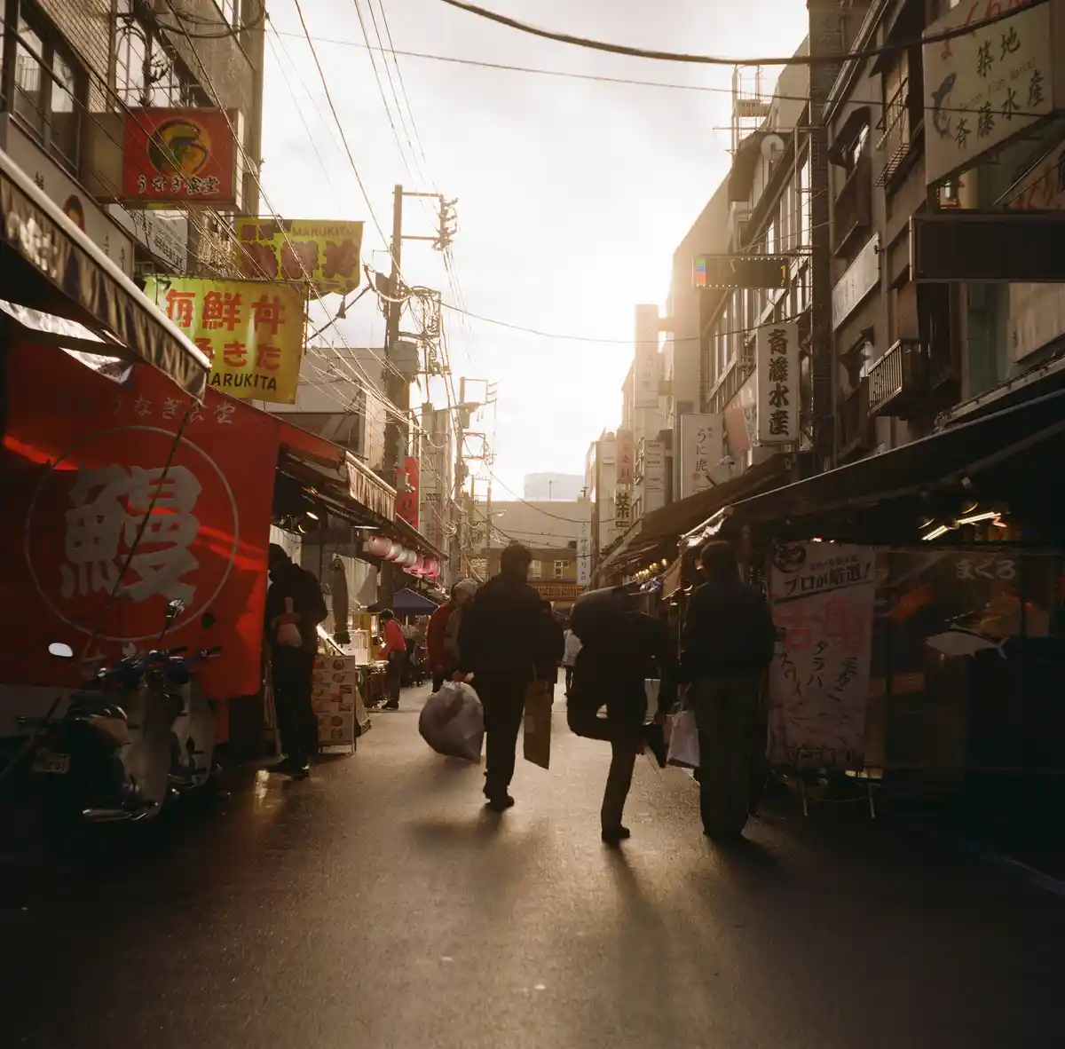 A photo of a bustling, narrow Japanese street market with people walking away from the camera, lined with colorful signs and…