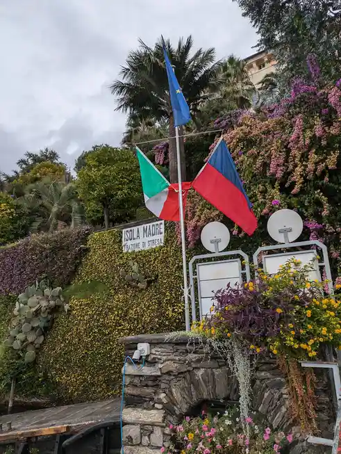 A photo of a lush green hillside with a sign reading 'ISOLA MADRE FRAZIONE DI STRESA' and three flags: the EU flag, the…