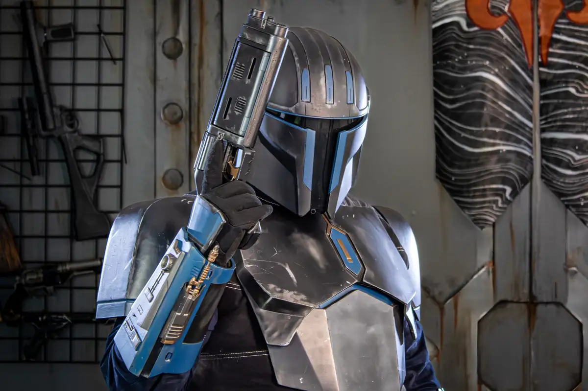 A person in a Mandalorian armor costume, featuring a helmet with blue accents and a metallic chest plate, holds a blaster…