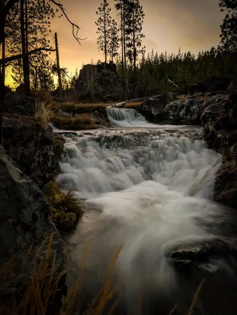 A long-exposure photograph of a cascading waterfall surrounded by dark pine trees and rocks under a golden sunset sky, with…