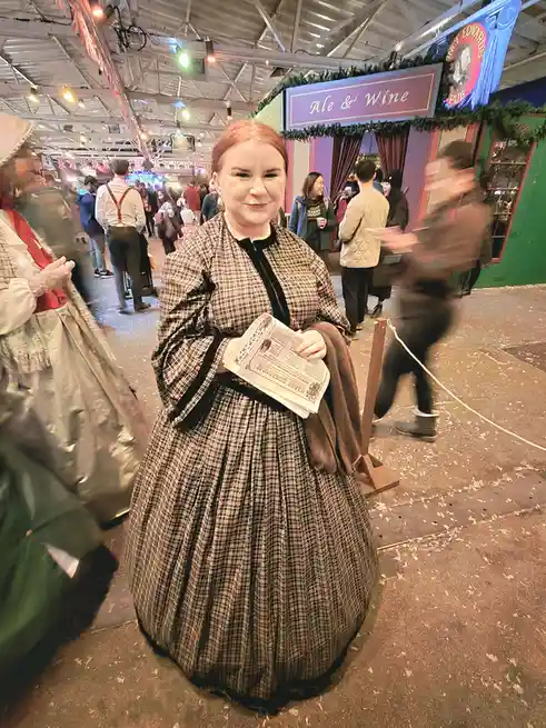 A woman in a historical-style plaid dress stands in a crowded indoor market or festival, holding a pamphlet. Behind her is a…