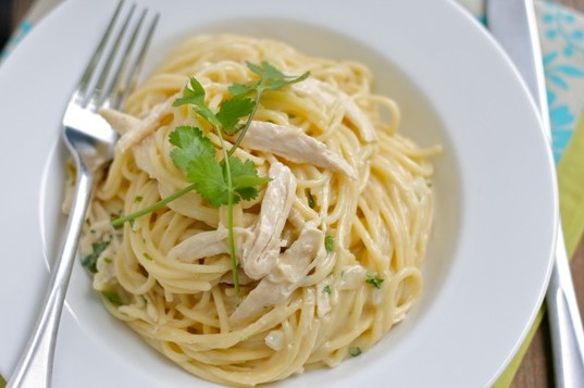 A close-up photo of a white bowl filled with creamy pasta mixed with shredded chicken and garnished with fresh cilantro. A…