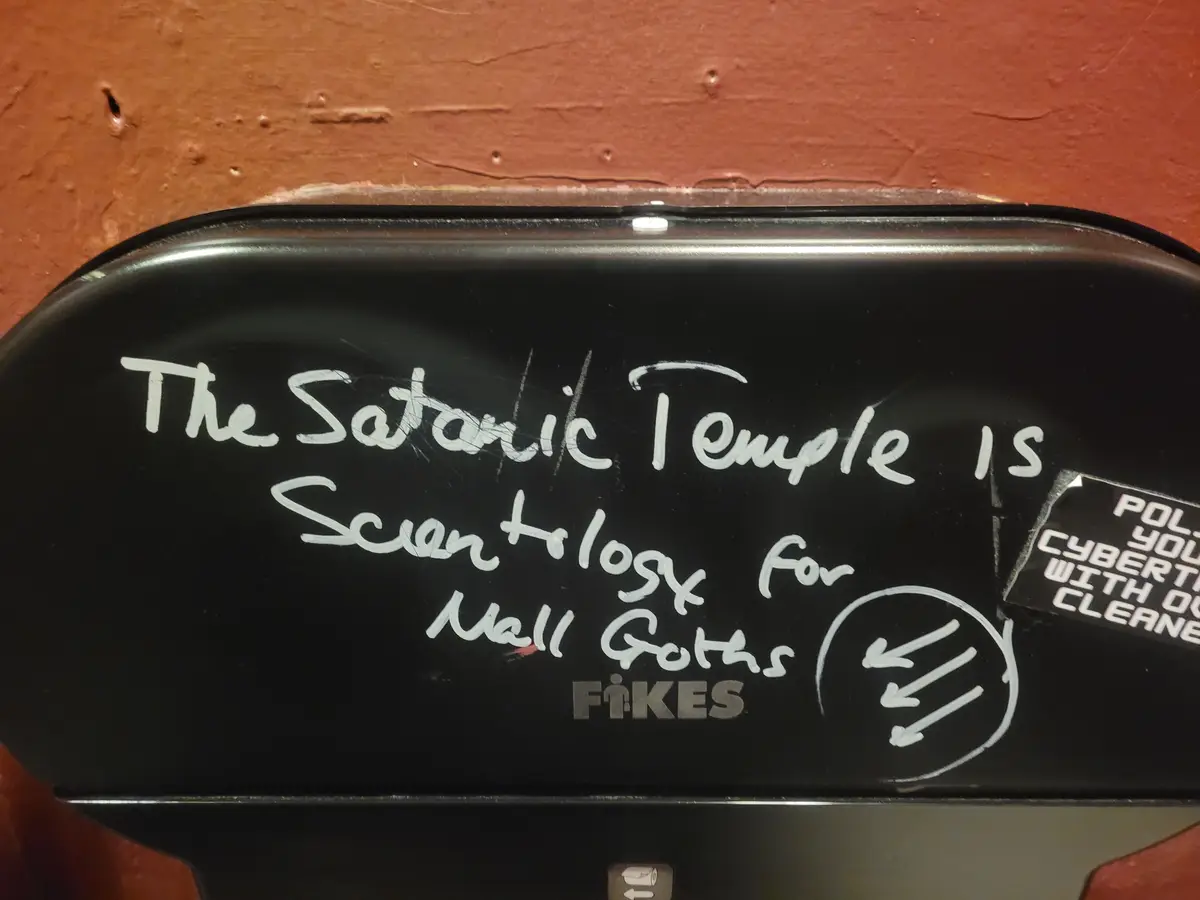 A close-up of a black public trash can with white handwritten text that reads 'The Satanic Temple is Scientology for Mall…