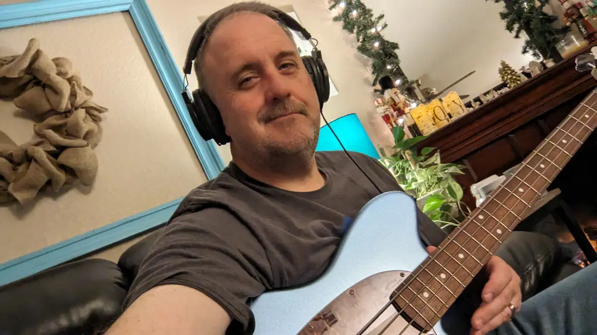 A man with headphones and a light blue bass guitar sits on a couch in a room decorated with Christmas lights, a wreath, and…