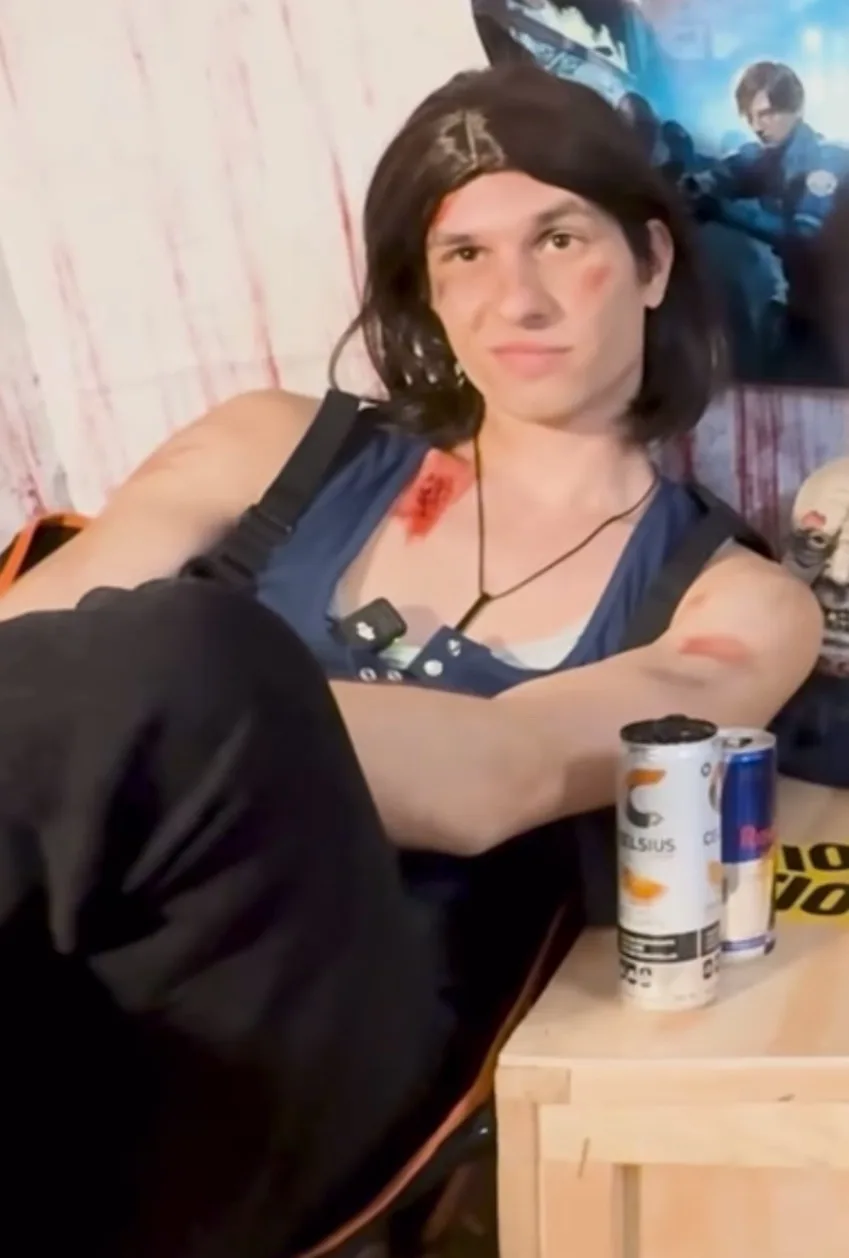 A person with a dark wig and fake blood on their chest and arm sits casually in a chair, looking relaxed. Two cans of…