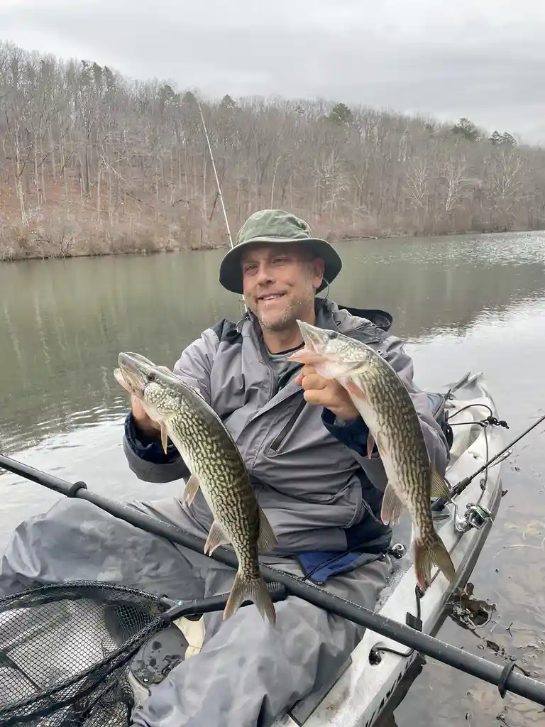 A man in a kayak on a calm river, wearing a green hat and gray fishing gear, proudly holds up two large pike fish, one in…