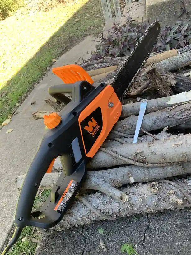 An orange and black WEN brand electric chainsaw rests on a pile of logs outdoors, with a white zip tie attached to one of…