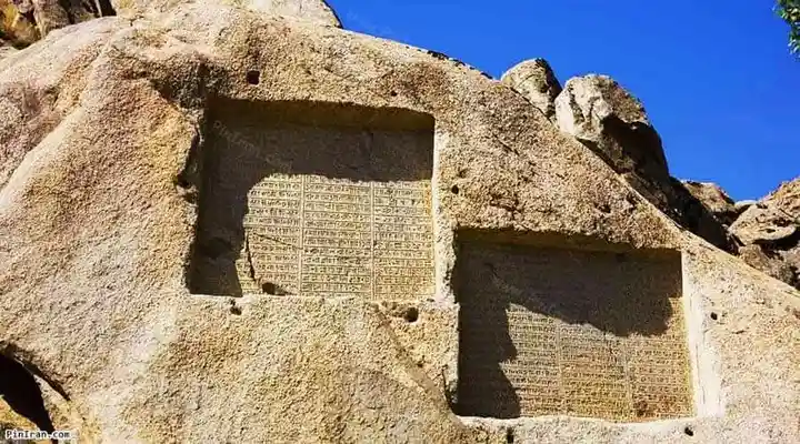 A large, sunlit rock face features two carved inscriptions; the left one is clearly legible with dense text, while the right…