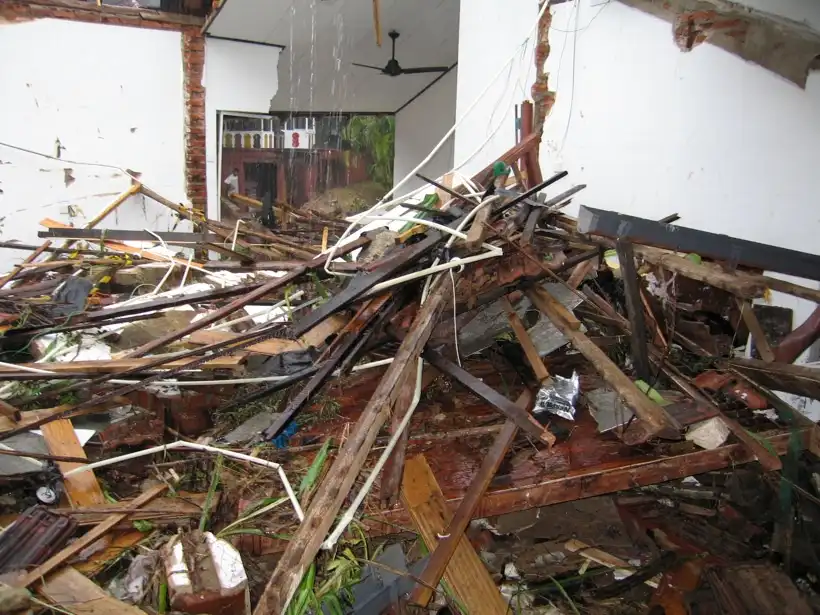 A room is in complete disarray with debris, broken wood, and scattered items, suggesting a collapse or severe damage. Water…