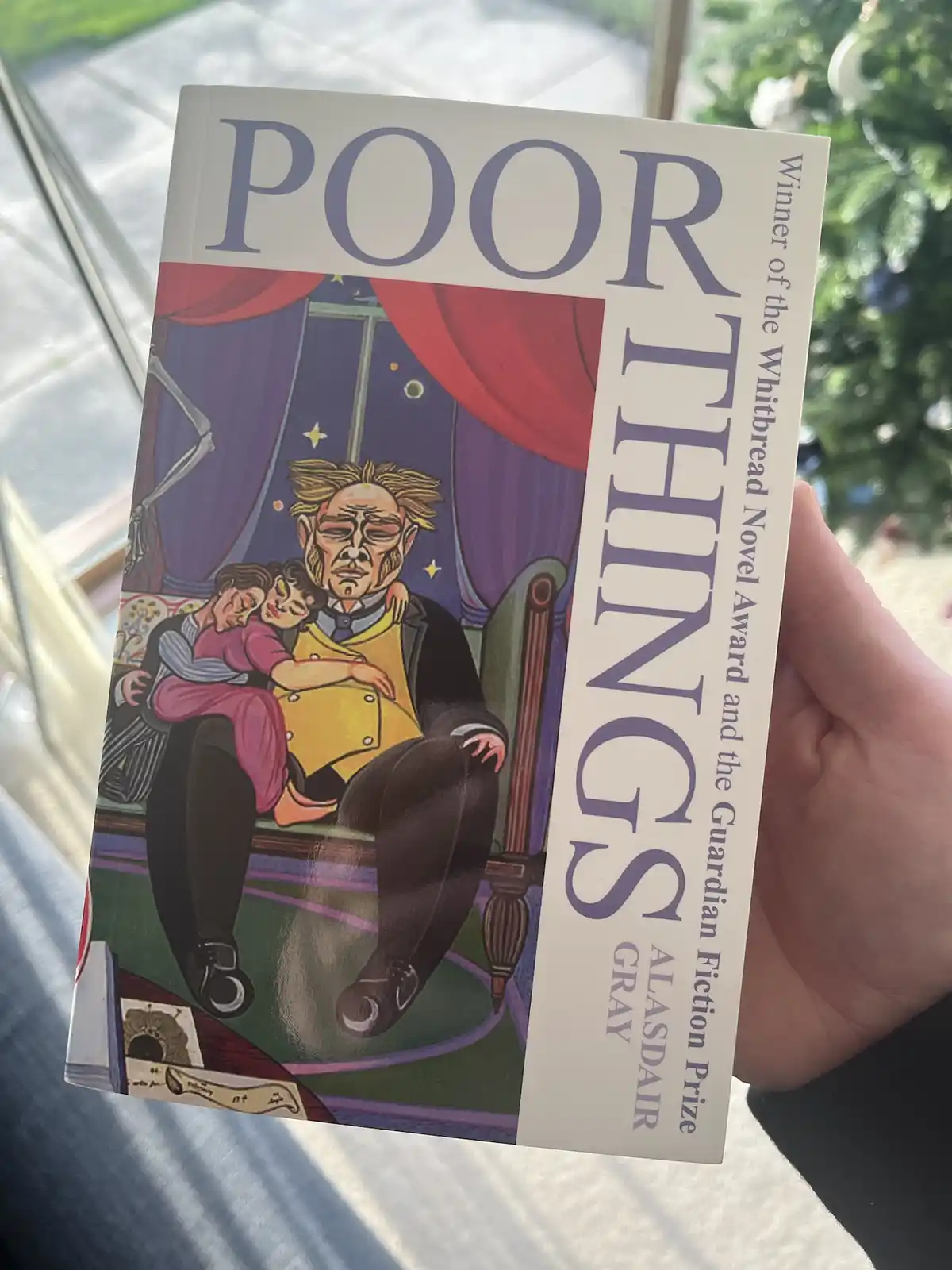 A hand holds a physical copy of the novel 'Poor Things' by Alasdair Gray, featuring a cover illustration of a man in a…
