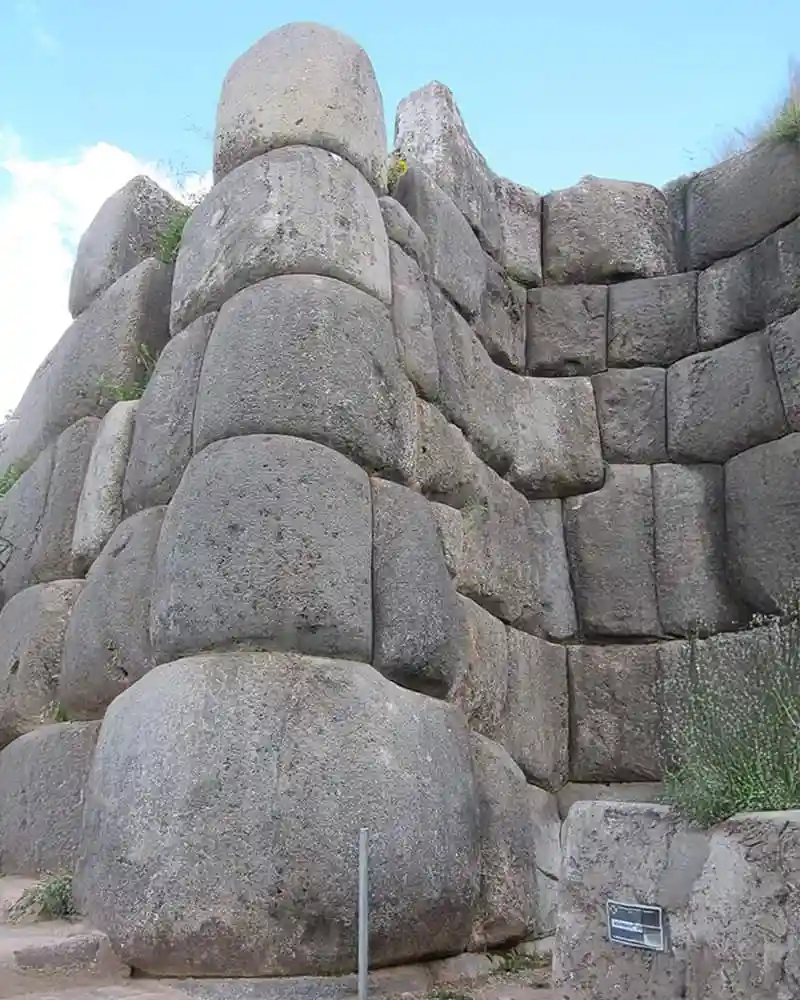 a low-angle photo of a massive ancient stone wall constructed from enormous, precisely fitted blocks of gray stone under a…
