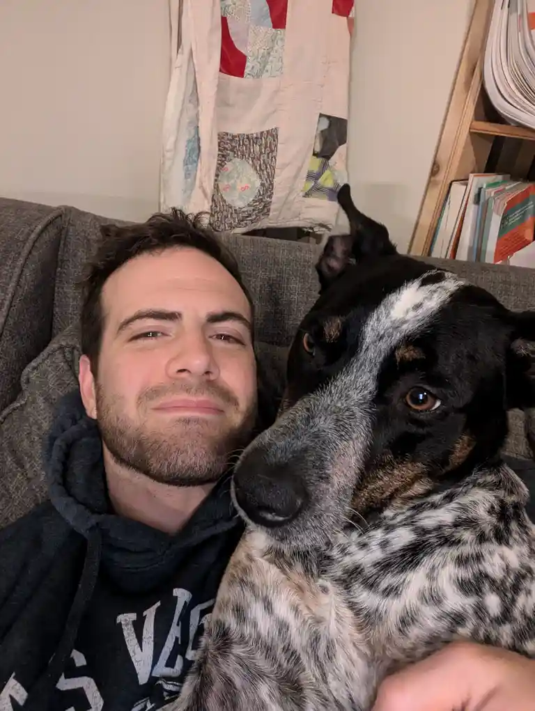 A man with a beard and dark hoodie sits on a grey couch next to a black and white speckled dog, both looking at the camera…