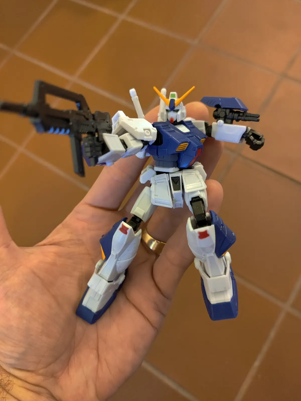 A small blue and white plastic robot toy, resembling a Gundam, is held in a person's hand against a tiled floor background…