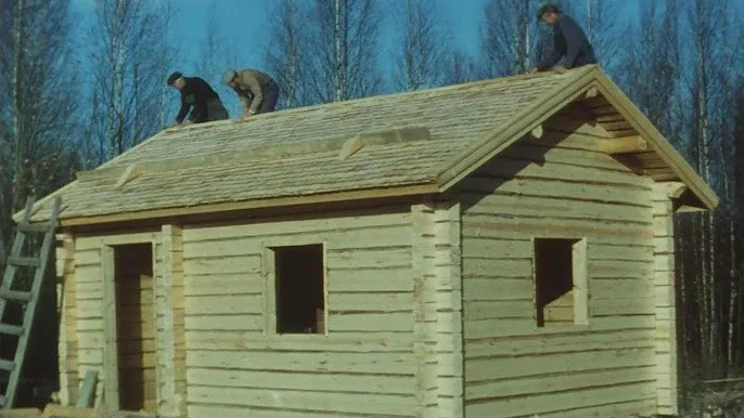 Three men are on the roof of a wooden cabin, installing shingles, while the structure is still unfinished with bare window…