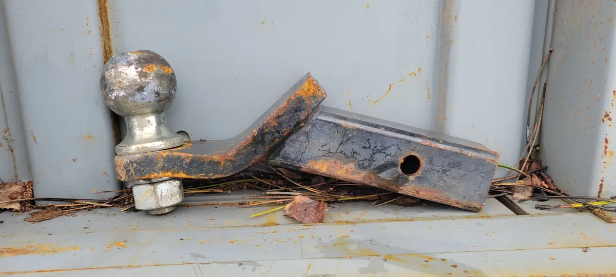 A rusty, weathered trailer hitch with a spherical ball mount is lying on its side against a gray metal surface, surrounded…