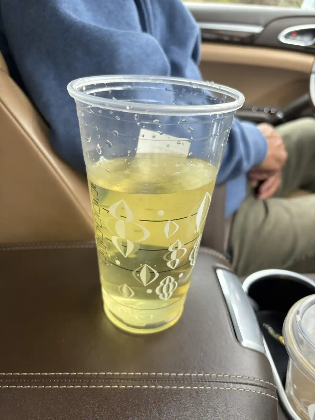 A clear plastic Starbucks cup filled with a pale yellow liquid sits on a car's center console, condensation beading on its…