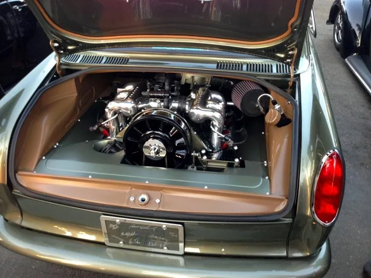 The image shows the rear of a vintage green Mini car with its trunk open, revealing a large, shiny, custom engine instead of…