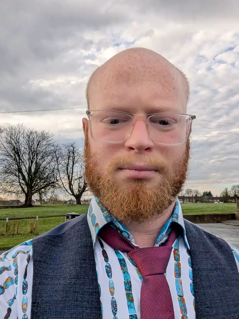 A close-up selfie of a bald, red-haired man with a full beard and clear-framed glasses, wearing a patterned shirt, a dark…