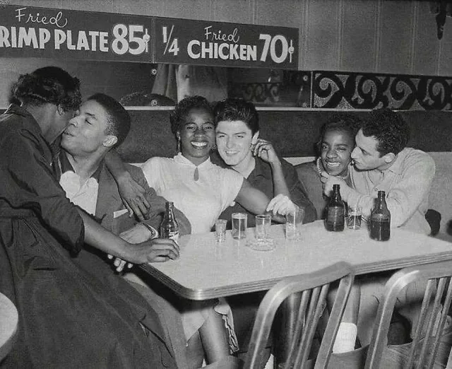 A black-and-white photo of six young people, mostly Black and one white man, sitting together at a diner table with beer…