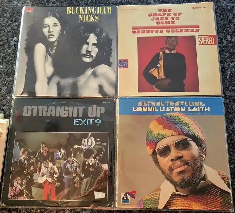 Four vintage vinyl record album covers are laid out on a dark countertop. Top left: Buckingham Nicks with a black-and-white…