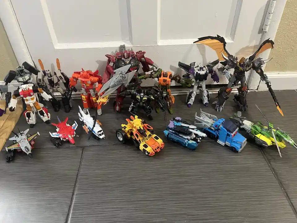 A collection of various Transformers action figures and vehicles are arranged on a dark gray wood-patterned floor in front…