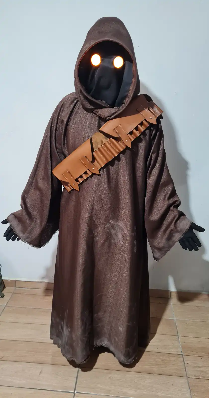A person in a brown hooded robe with glowing orange eyes and a tan leather strap across the chest stands against a plain…