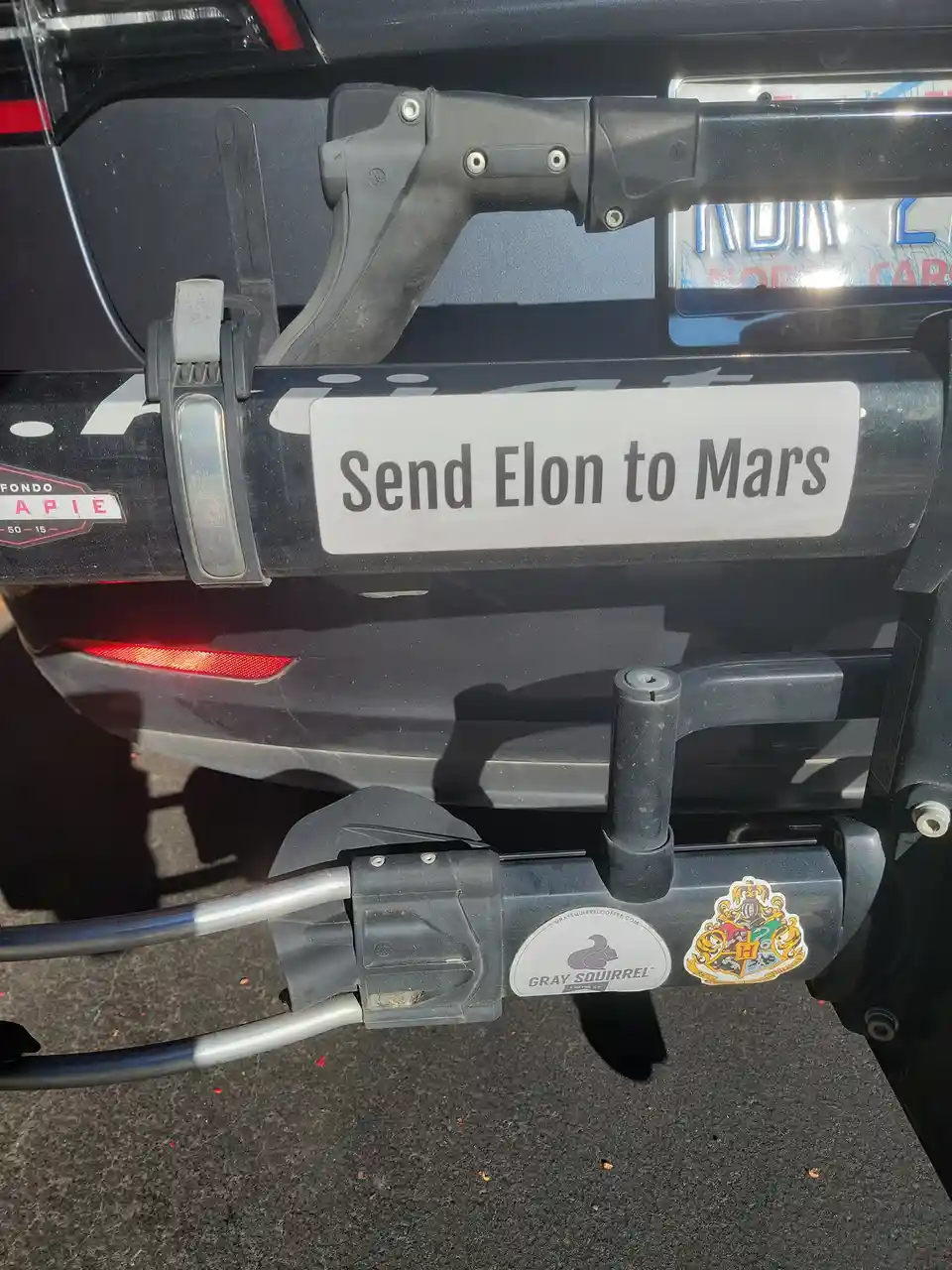 A close-up of the rear of a dark-colored vehicle showing a custom license plate holder with a white sticker that reads 'Send…