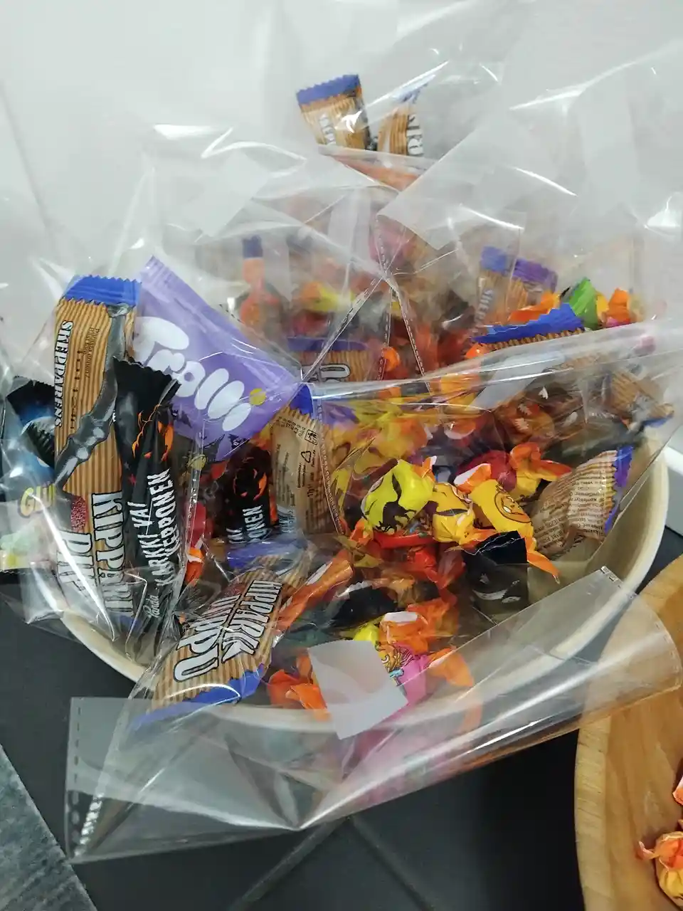 A white bowl is overflowing with Halloween-themed candy, including individually wrapped treats in orange, black, and purple…