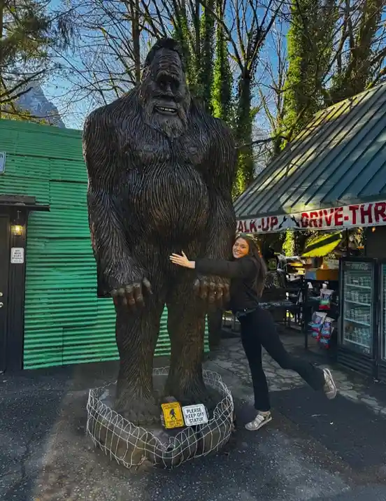 A woman poses playfully next to a large, dark brown statue of Bigfoot, smiling and leaning against its leg. The statue…