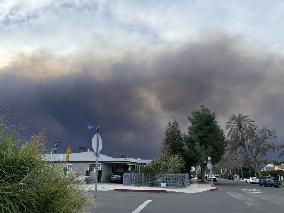 A residential street corner with houses, cars, and street signs under a massive, ominous dark smoke cloud that dominates the…