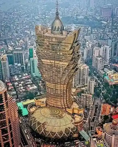 An aerial view of the Grand Lisboa hotel in Macau, a distinctive golden skyscraper with a flared, tiered design and a domed…
