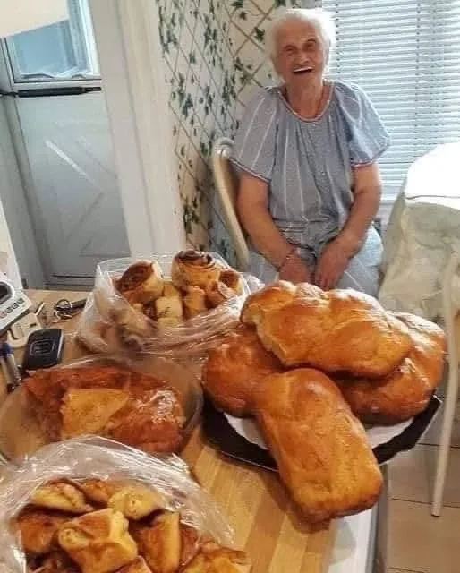 An elderly woman with white hair sits in a kitchen chair behind a wooden table overflowing with large loaves of bread and…