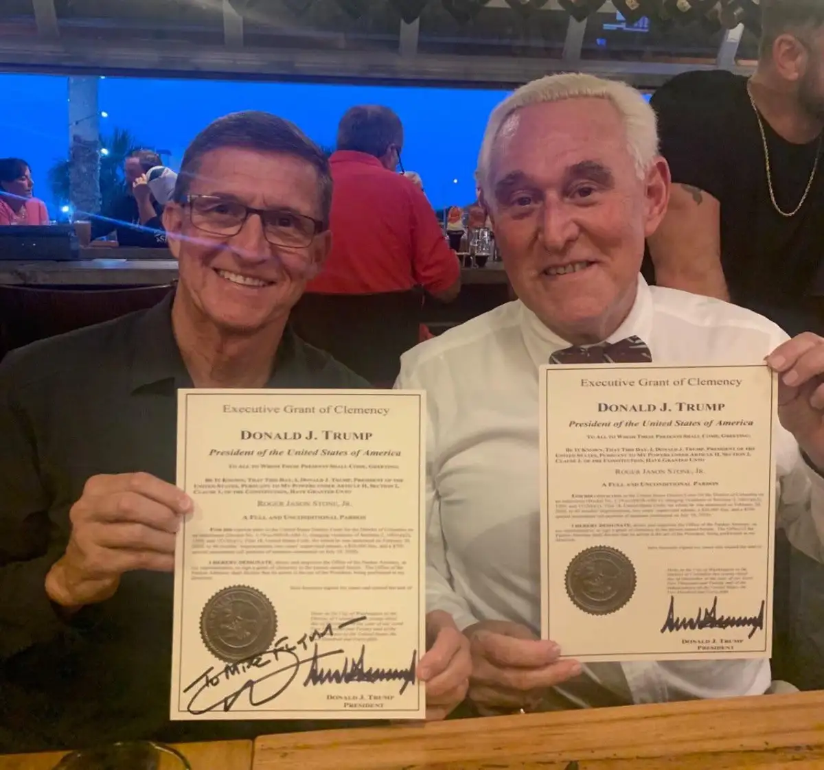 Two men are smiling and holding up identical fake pardons that appear to be signed by Donald J. Trump, granting a full and…