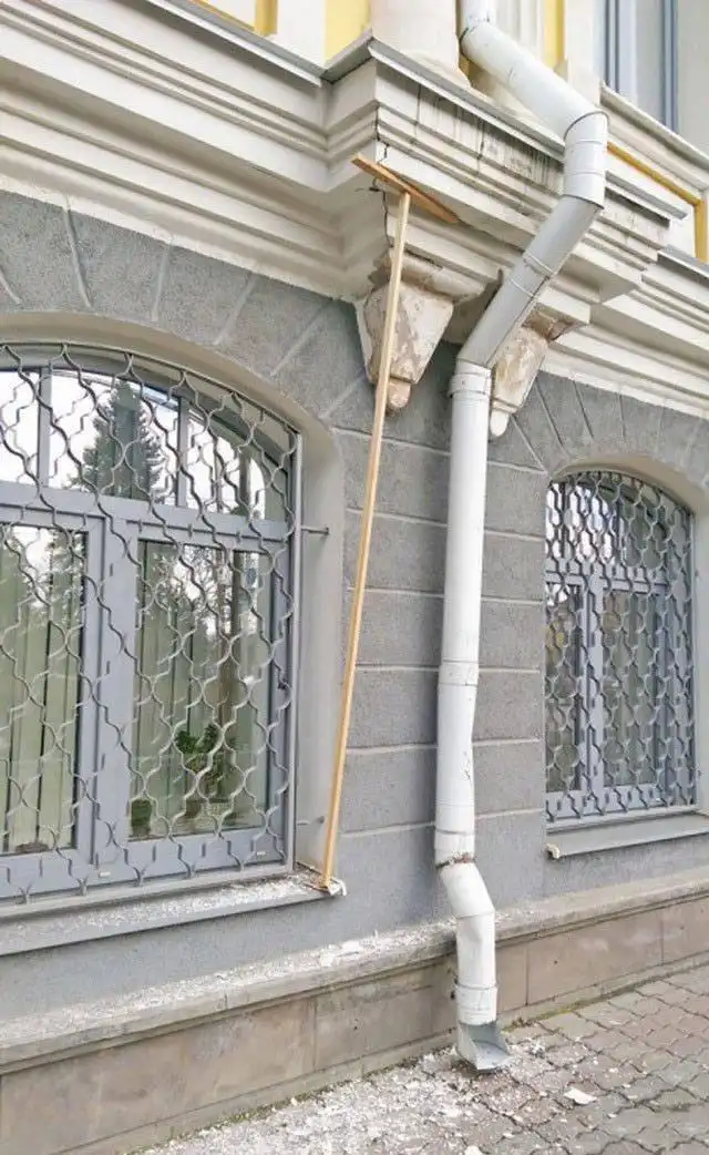 An exterior shot of a building with gray stone walls and arched windows with metal grilles. A long wooden broom is propped…