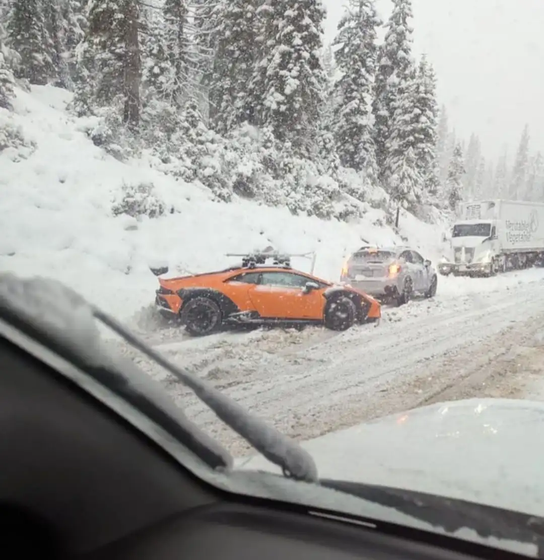 A bright orange Lamborghini is stuck sideways in deep snow on a snowy mountain road, viewed from inside another vehicle…