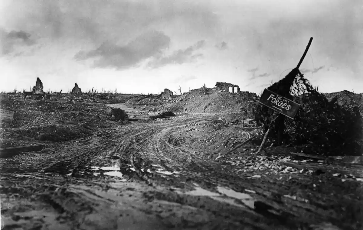 A black-and-white historical photograph shows a devastated, muddy battlefield with ruined buildings and debris under a…