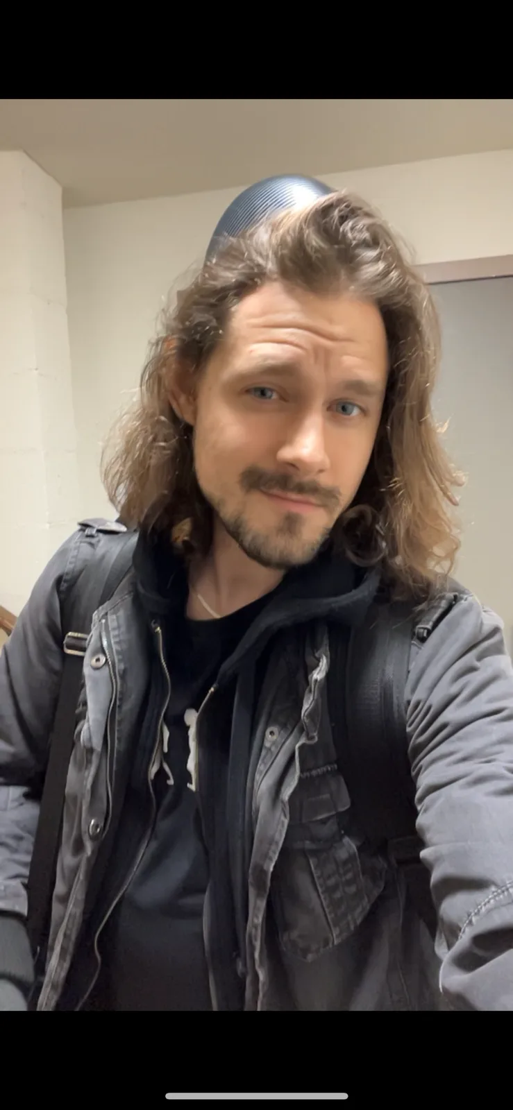 A man with long brown hair and a beard, wearing a dark jacket and backpack, looks at the camera with a skeptical expression…